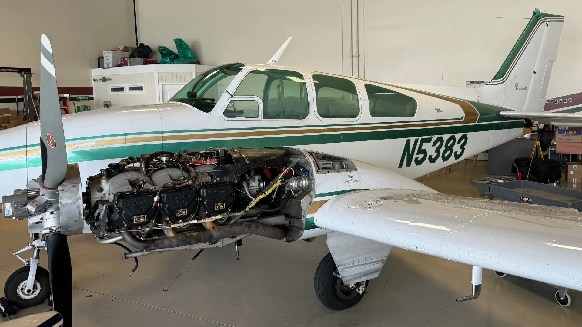 Beechcraft Baron in a maintenance hangar with the left engine cowling removed, exposing the cylinders, exhaust, and wiring